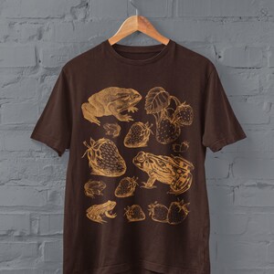 Pu&ograve; includere: Una maglietta marrone con una stampa dorata di rane e fragole. Il design presenta pi&ugrave; rane e fragole in stile vintage.