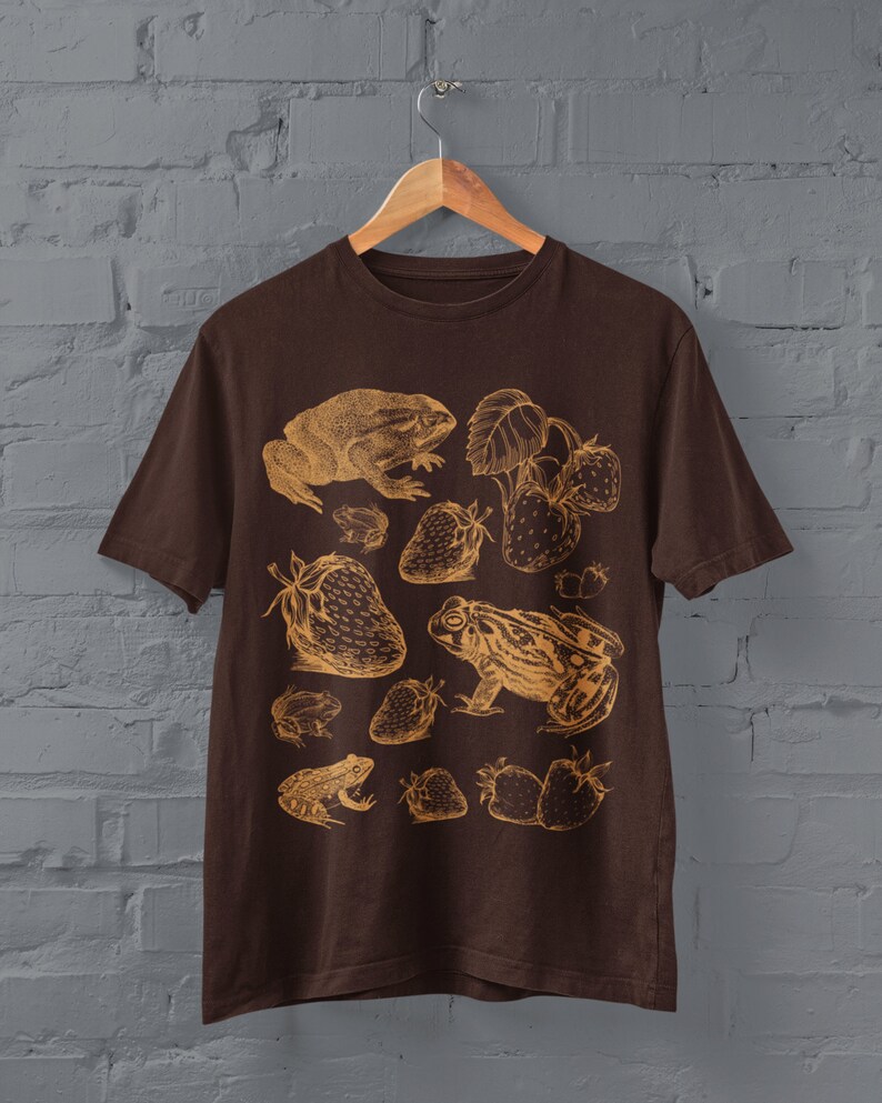 Pu&ograve; includere: Una maglietta marrone con una stampa dorata di rane e fragole. Il design presenta pi&ugrave; rane e fragole in stile vintage.