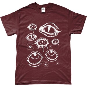 May include: Burgundy t-shirt featuring a white graphic of multiple stylized eyes. The eyes vary in size and style, some with teardrop details. The shirt is made of a soft material and has a classic crew neck.