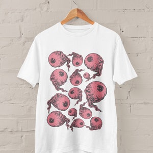 May include: White t-shirt with a graphic print of pink eyes with black outlines and smoke-like trails. The eyes are arranged in a random pattern on the shirt.