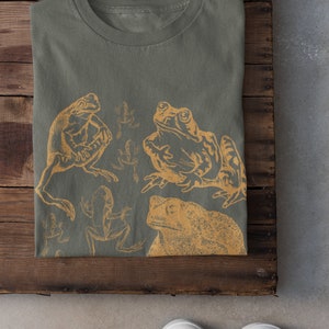 Pode incluir: Uma camiseta verde com uma estampa de v&aacute;rias r&atilde;s de cor amarela. As r&atilde;s est&atilde;o em diferentes poses e tamanhos. A camiseta est&aacute; dobrada e colocada sobre uma superf&iacute;cie de madeira.