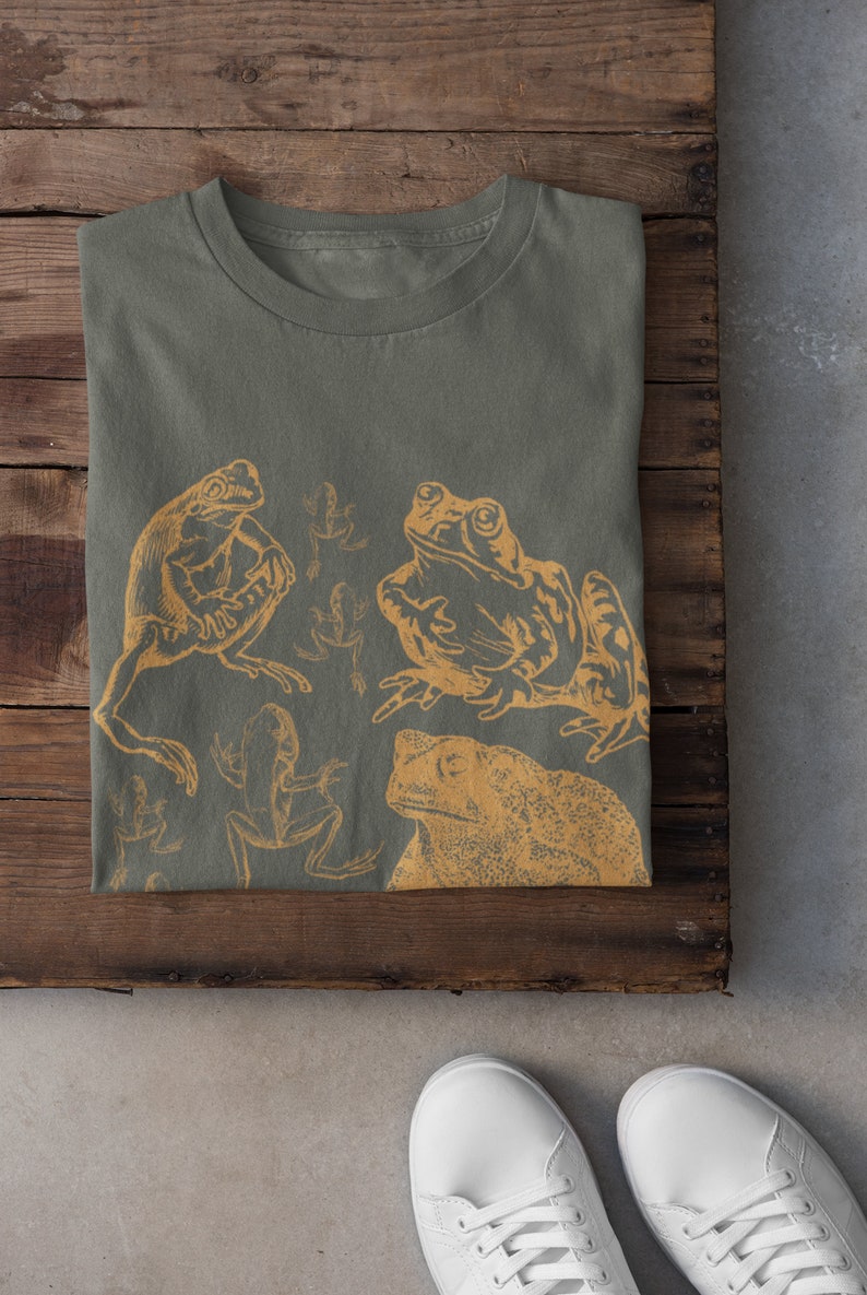 Pode incluir: Uma camiseta verde com uma estampa de v&aacute;rias r&atilde;s de cor amarela. As r&atilde;s est&atilde;o em diferentes poses e tamanhos. A camiseta est&aacute; dobrada e colocada sobre uma superf&iacute;cie de madeira.
