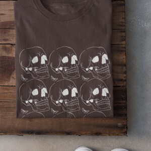 May include: A brown t-shirt with a white skull and butterfly pattern. The shirt is folded and laid on a wooden surface.
