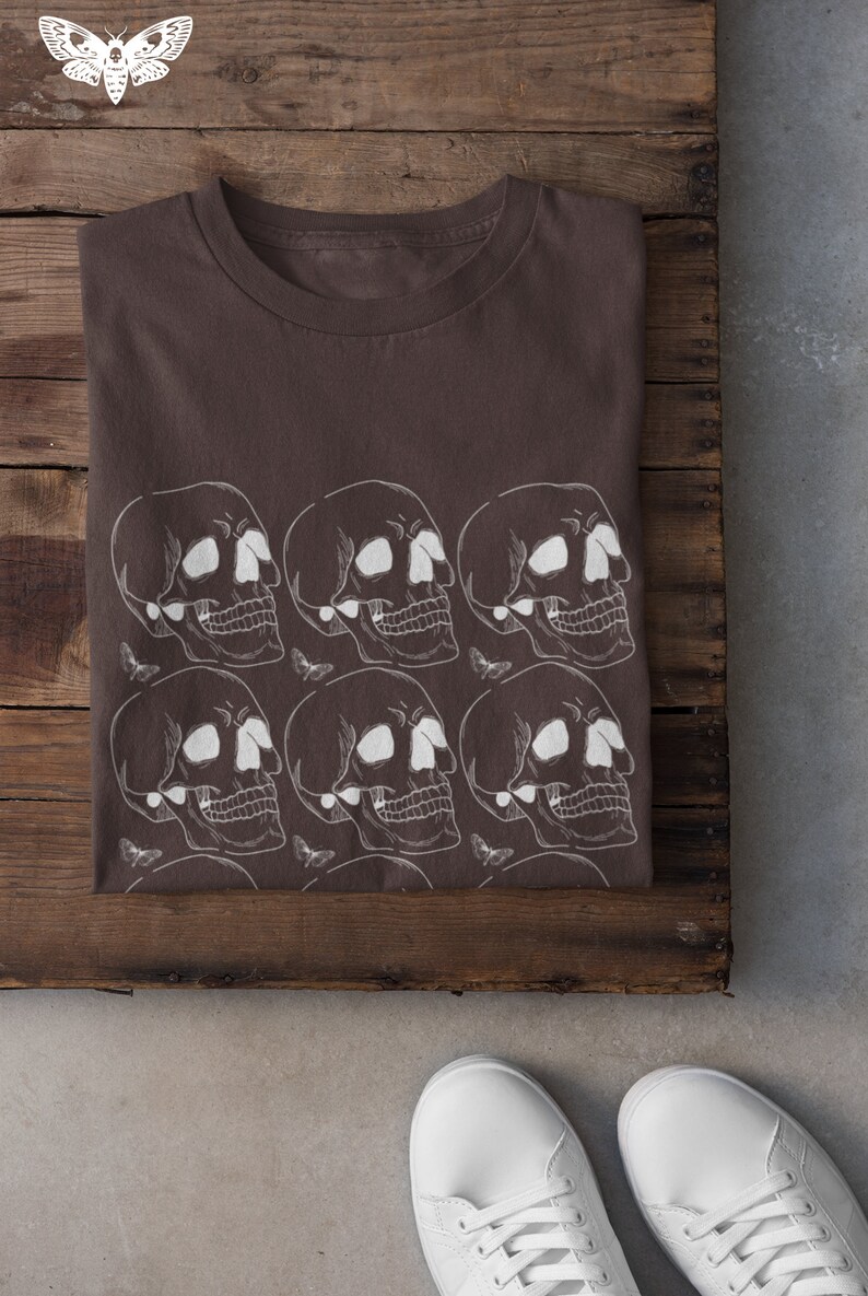 May include: A brown t-shirt with a white skull and butterfly pattern. The shirt is folded and laid on a wooden surface.