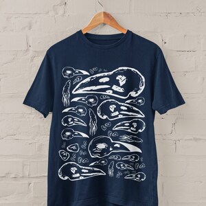 Op de afbeelding: Marineblauw t-shirt met een witte print van vogel schedels in verschillende maten en ori&euml;ntaties. Het ontwerp cre&euml;ert een herhalend patroon. Het t-shirt hangt aan een houten hanger tegen een witte bakstenen muur.