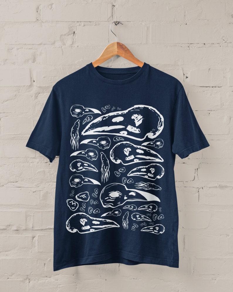 Op de afbeelding: Marineblauw t-shirt met een witte print van vogel schedels in verschillende maten en ori&euml;ntaties. Het ontwerp cre&euml;ert een herhalend patroon. Het t-shirt hangt aan een houten hanger tegen een witte bakstenen muur.