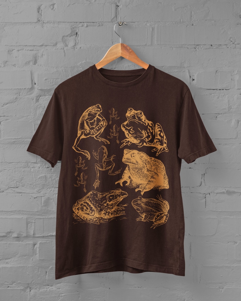 Pode incluir: Uma camiseta marrom com uma estampa gr&aacute;fica de v&aacute;rios sapos e r&atilde;s em uma cor marrom claro. O design apresenta uma variedade de poses de sapos, incluindo sentados, pulando e nadando.