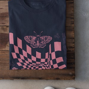 May include: A navy blue t-shirt with a pink and black checkered pattern, a butterfly graphic, and stars. The shirt is folded and laid on a wooden surface.