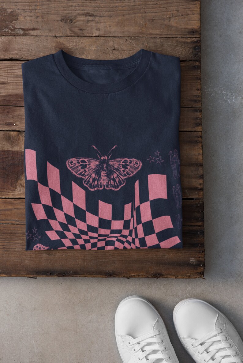 May include: A navy blue t-shirt with a pink and black checkered pattern, a butterfly graphic, and stars. The shirt is folded and laid on a wooden surface.