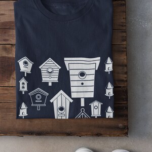 May include: A navy blue t-shirt featuring a white design of various birdhouses. The birdhouses are of different shapes and sizes, and some have small details like windows and perches.