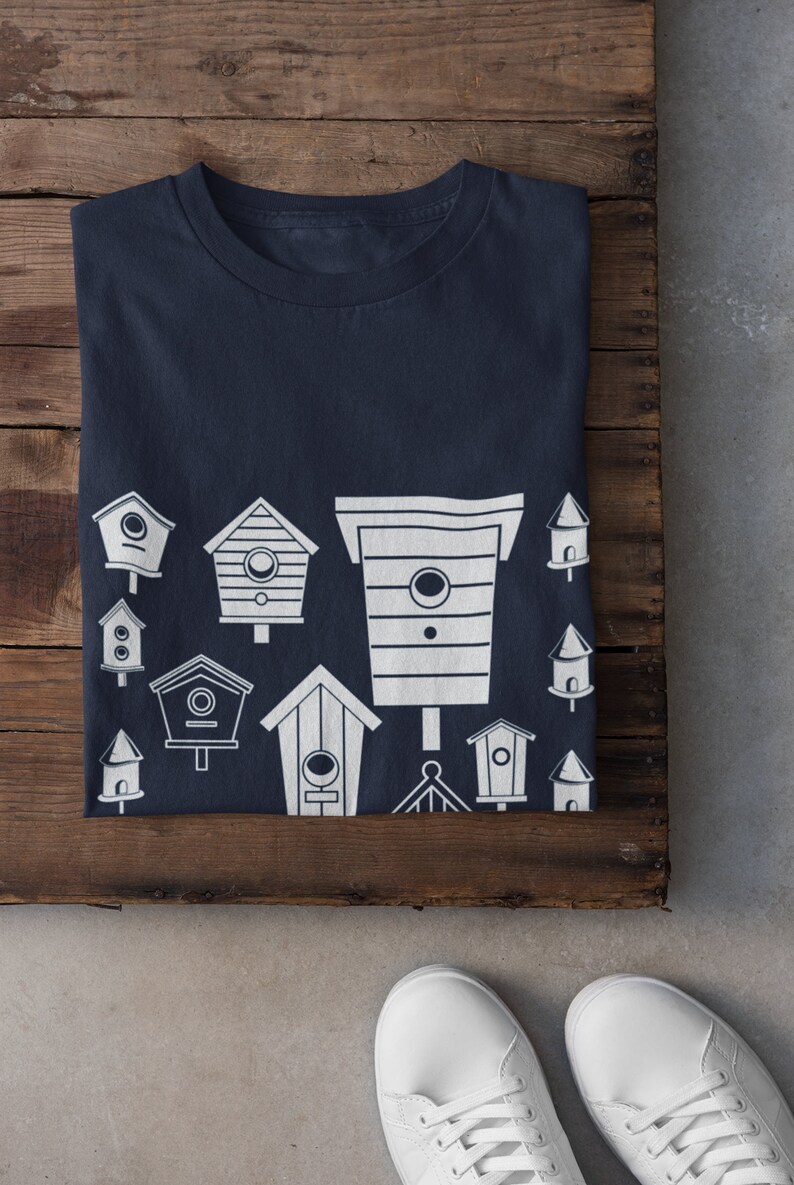 May include: A navy blue t-shirt featuring a white design of various birdhouses. The birdhouses are of different shapes and sizes, and some have small details like windows and perches.