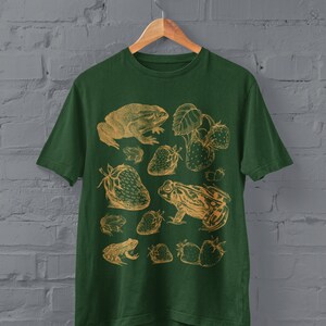 Pu&ograve; includere: Una maglietta verde con un'illustrazione vintage di rane e fragole in color oro. Il design presenta diverse rane e fragole in un motivo ripetitivo.