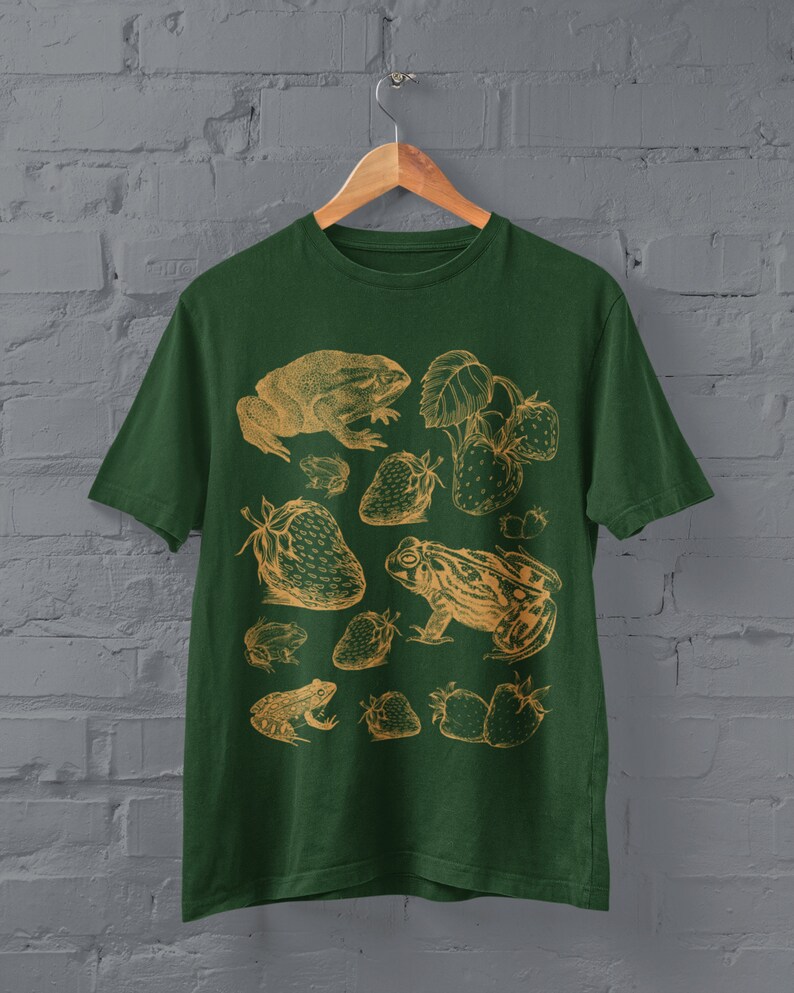 Pu&ograve; includere: Una maglietta verde con un'illustrazione vintage di rane e fragole in color oro. Il design presenta diverse rane e fragole in un motivo ripetitivo.