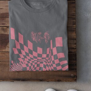 May include: A grey t-shirt with a pink checkered pattern and a graphic of three figures in a distorted perspective.