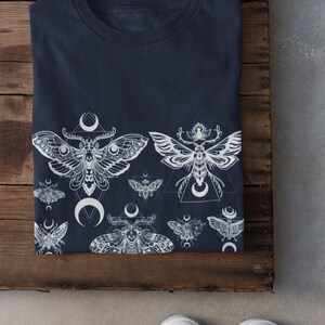 May include: A navy blue t-shirt with a white print of moths and crescent moons. The moths are stylized and have intricate details. The design is repeated across the bottom of the shirt.