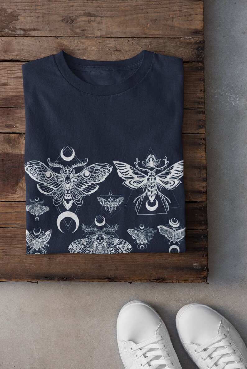 May include: A navy blue t-shirt with a white print of moths and crescent moons. The moths are stylized and have intricate details. The design is repeated across the bottom of the shirt.