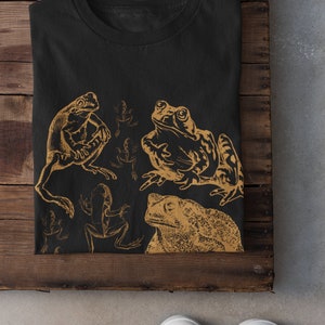 Pode incluir: Camiseta preta com uma estampa dourada de v&aacute;rios sapos e r&atilde;s. A estampa apresenta um sapo grande no centro, uma r&atilde; menor &agrave; esquerda e v&aacute;rias r&atilde;s menores no fundo.