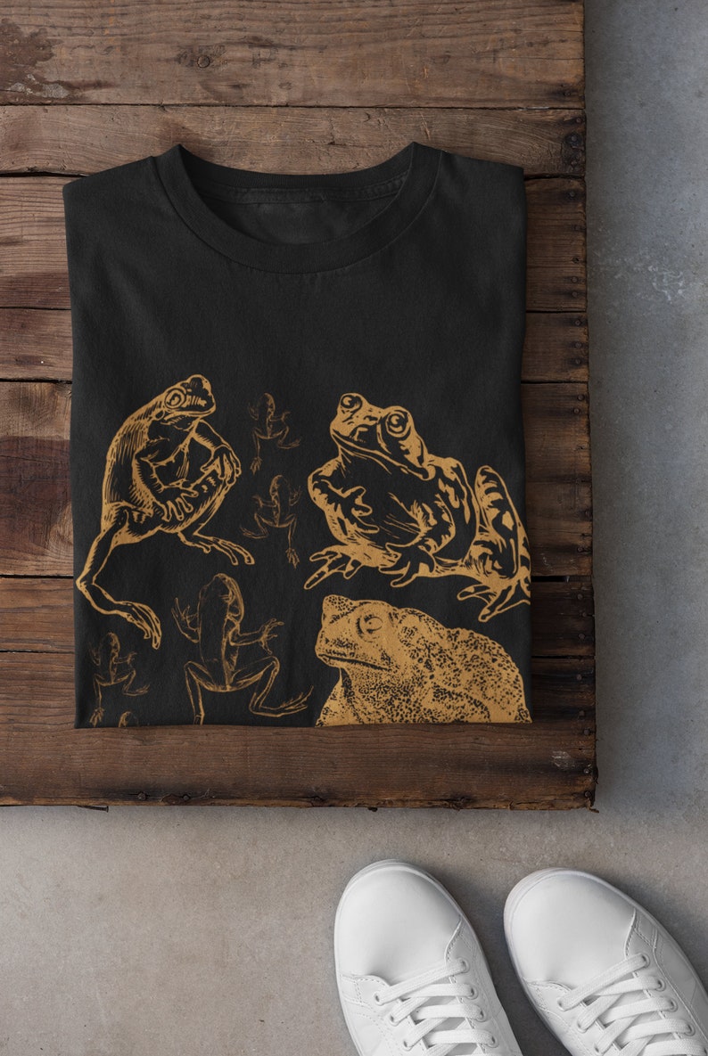 Pode incluir: Camiseta preta com uma estampa dourada de v&aacute;rios sapos e r&atilde;s. A estampa apresenta um sapo grande no centro, uma r&atilde; menor &agrave; esquerda e v&aacute;rias r&atilde;s menores no fundo.