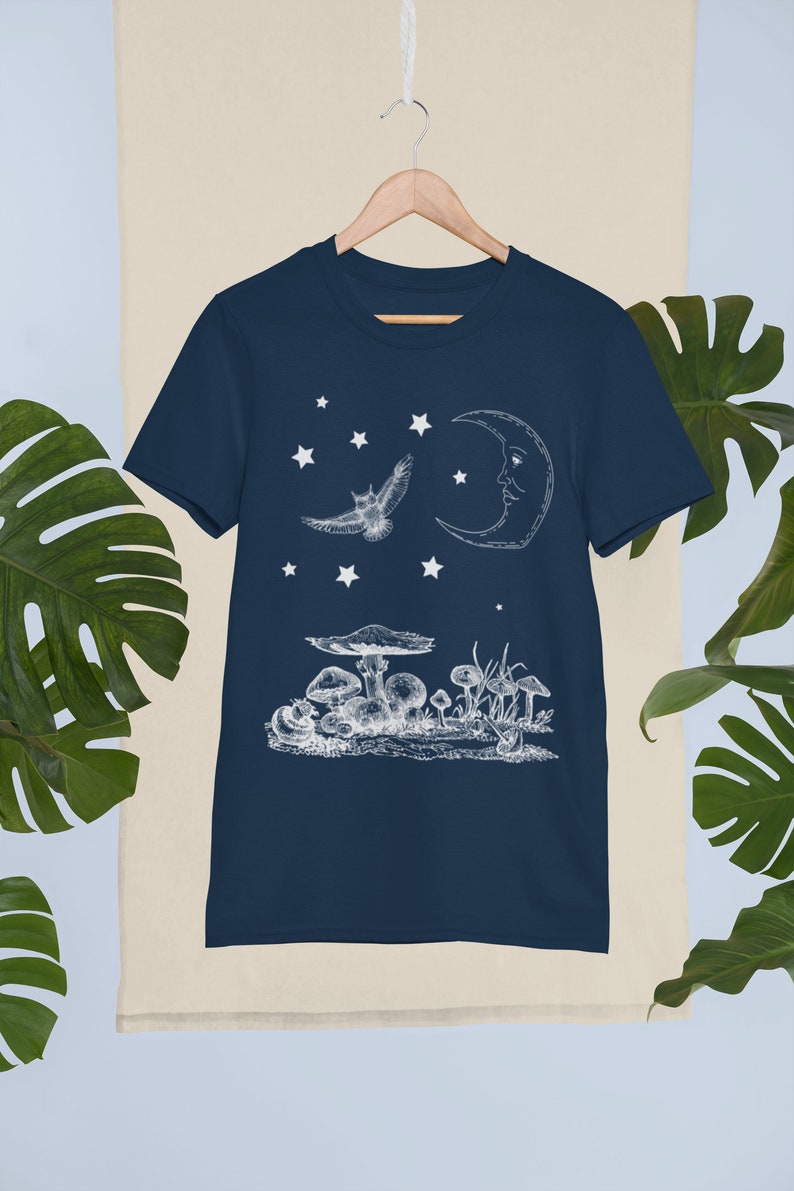 May include: A navy blue t-shirt with a white graphic of a crescent moon, an owl flying, and mushrooms. The graphic is reminiscent of a vintage illustration.