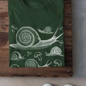 May include: A dark green t-shirt with a white print of snails and mushrooms. The snails are drawn in a vintage style.