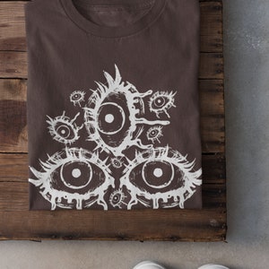 May include: A brown t-shirt with a white graphic print of multiple eyes. The eyes are stylized and have a dripping effect. The shirt is folded and laid on a wooden surface.