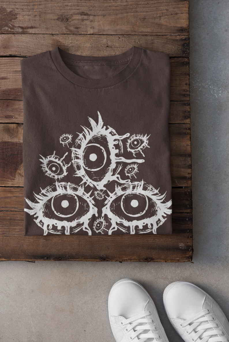 May include: A brown t-shirt with a white graphic print of multiple eyes. The eyes are stylized and have a dripping effect. The shirt is folded and laid on a wooden surface.