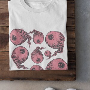May include: White t-shirt with a pink and black eye print design. The eyes are stylized and have a spooky, surreal look.