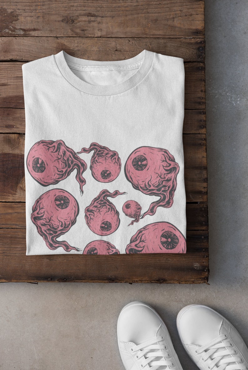 May include: White t-shirt with a pink and black eye print design. The eyes are stylized and have a spooky, surreal look.