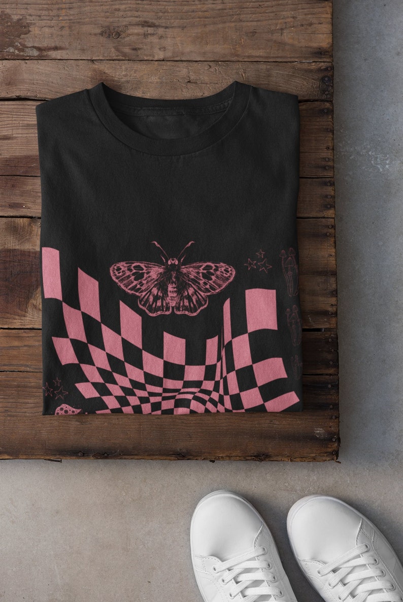 May include: Black t-shirt with a pink checkered pattern and a butterfly graphic. The pattern is a distorted checkerboard with a pink butterfly in the center.
