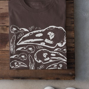 Op de afbeelding: Een gevouwen bruin t-shirt met een wit grafisch ontwerp met vogel schedels en abstracte vormen. Het shirt wordt gepresenteerd op een houten oppervlak, met witte sneakers onderaan.