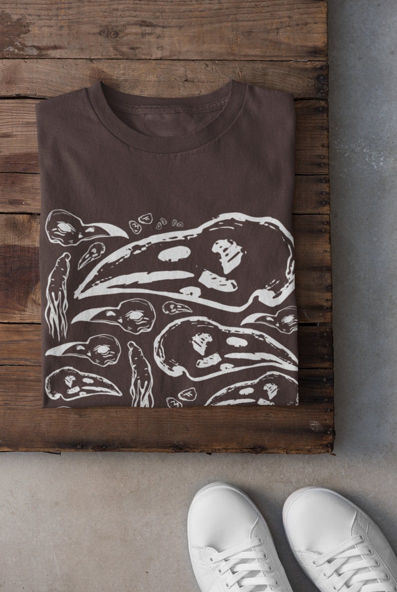Op de afbeelding: Een gevouwen bruin t-shirt met een wit grafisch ontwerp met vogel schedels en abstracte vormen. Het shirt wordt gepresenteerd op een houten oppervlak, met witte sneakers onderaan.
