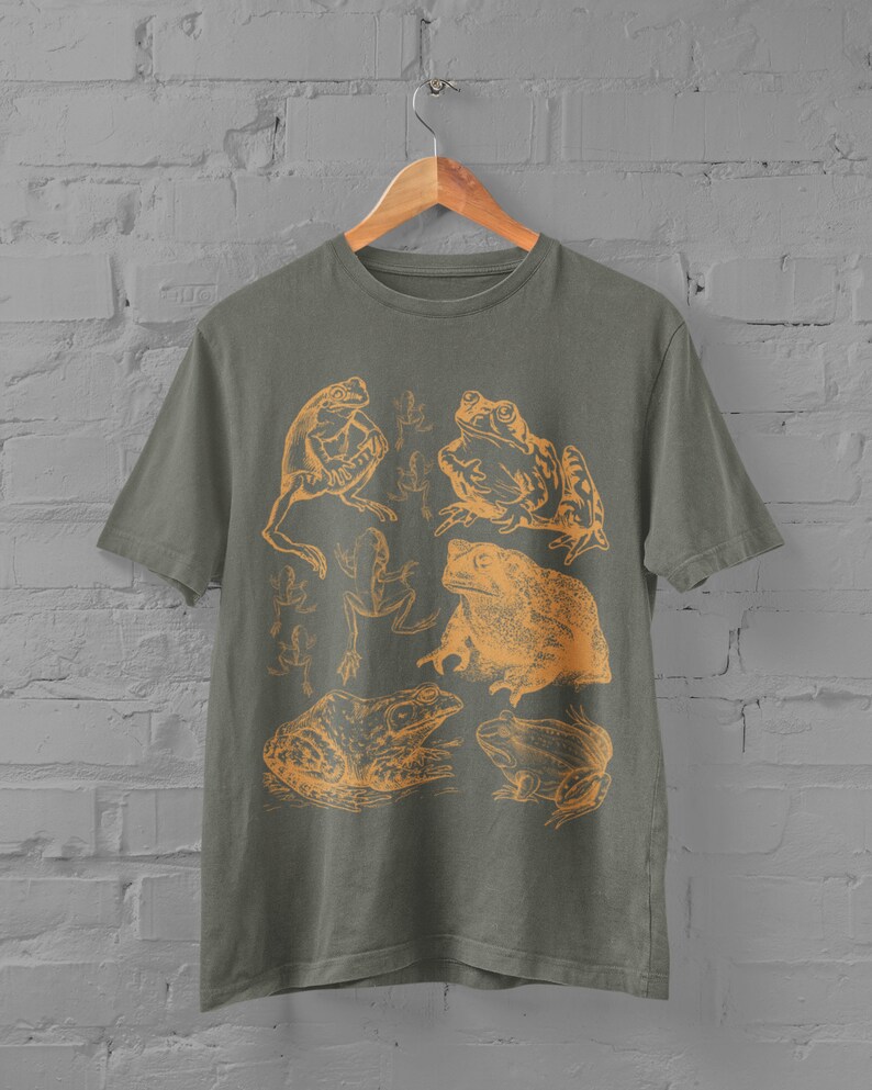 Pode incluir: Uma camiseta verde com uma estampa laranja de v&aacute;rios sapos e r&atilde;s. A estampa inclui um sapo com textura irregular, uma r&atilde; com l&iacute;ngua longa e uma r&atilde; com cabe&ccedil;a grande.