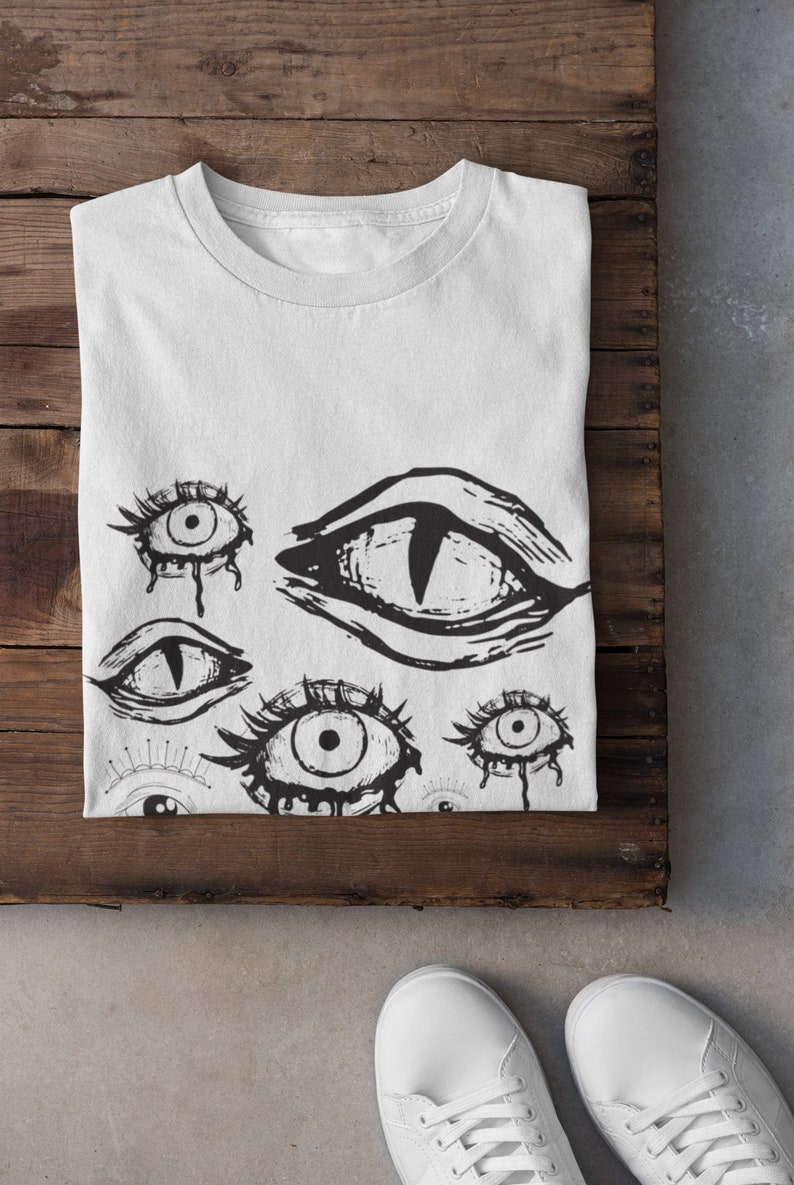 May include: White t-shirt with a black and white graphic print of multiple stylized eyes. The eyes are drawn in a creepy, abstract style.