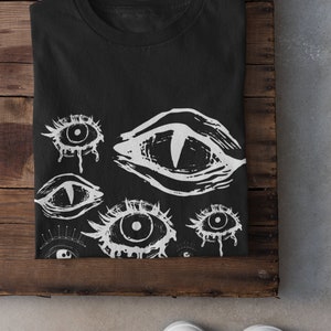 May include: Black t-shirt with a white print of multiple stylized eyes. The eyes are drawn in a graphic style with dripping lines.