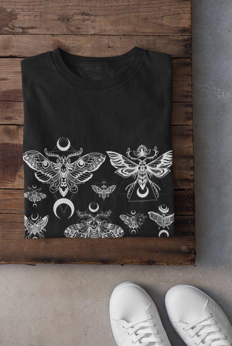 May include: Black t-shirt with a white print of moths and crescent moons. The moths are stylized and have intricate details. The design is repeated across the bottom of the shirt.