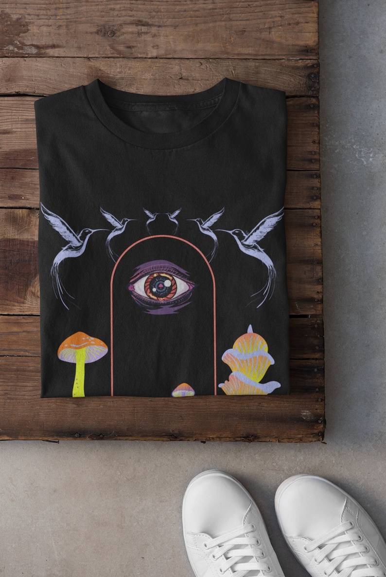 以下が含まれることがあります： 大きな目、キノコ、鳥が描かれたサイケデリックなデザインの黒いTシャツ。目はピンクとオレンジのアーチで囲まれています。鳥は目の周りを円を描くように飛んでいます。