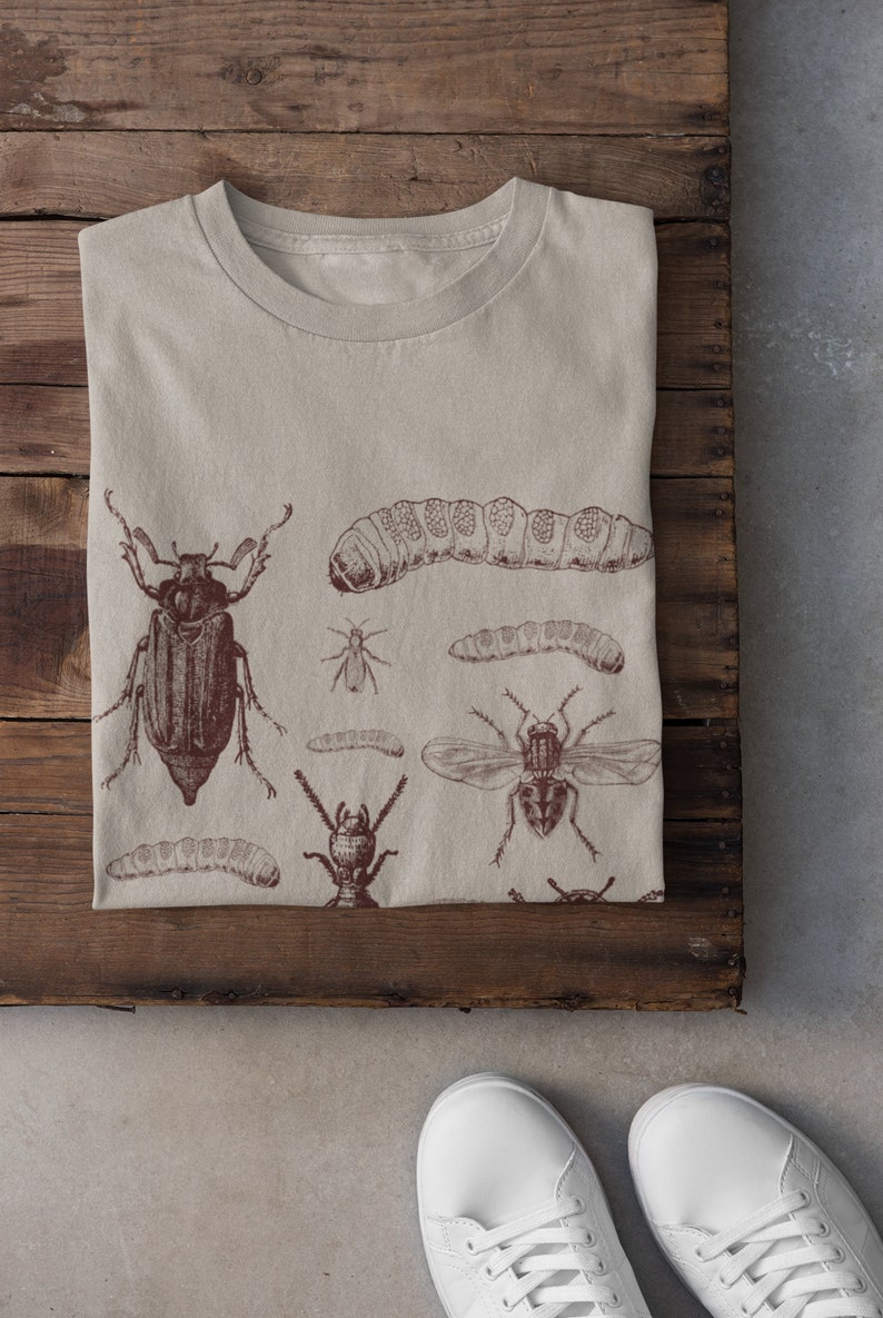 May include: Beige t-shirt with detailed dark brown illustrations of insects, including beetles, caterpillars, and flies. The shirt is folded on a wooden surface. White trainers are visible at the bottom of the image.