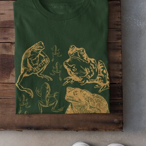 Pode incluir: Uma camiseta verde com uma estampa dourada de sapos e r&atilde;s. A estampa apresenta uma variedade de poses de sapos, incluindo um sapo grande com um olho proeminente.