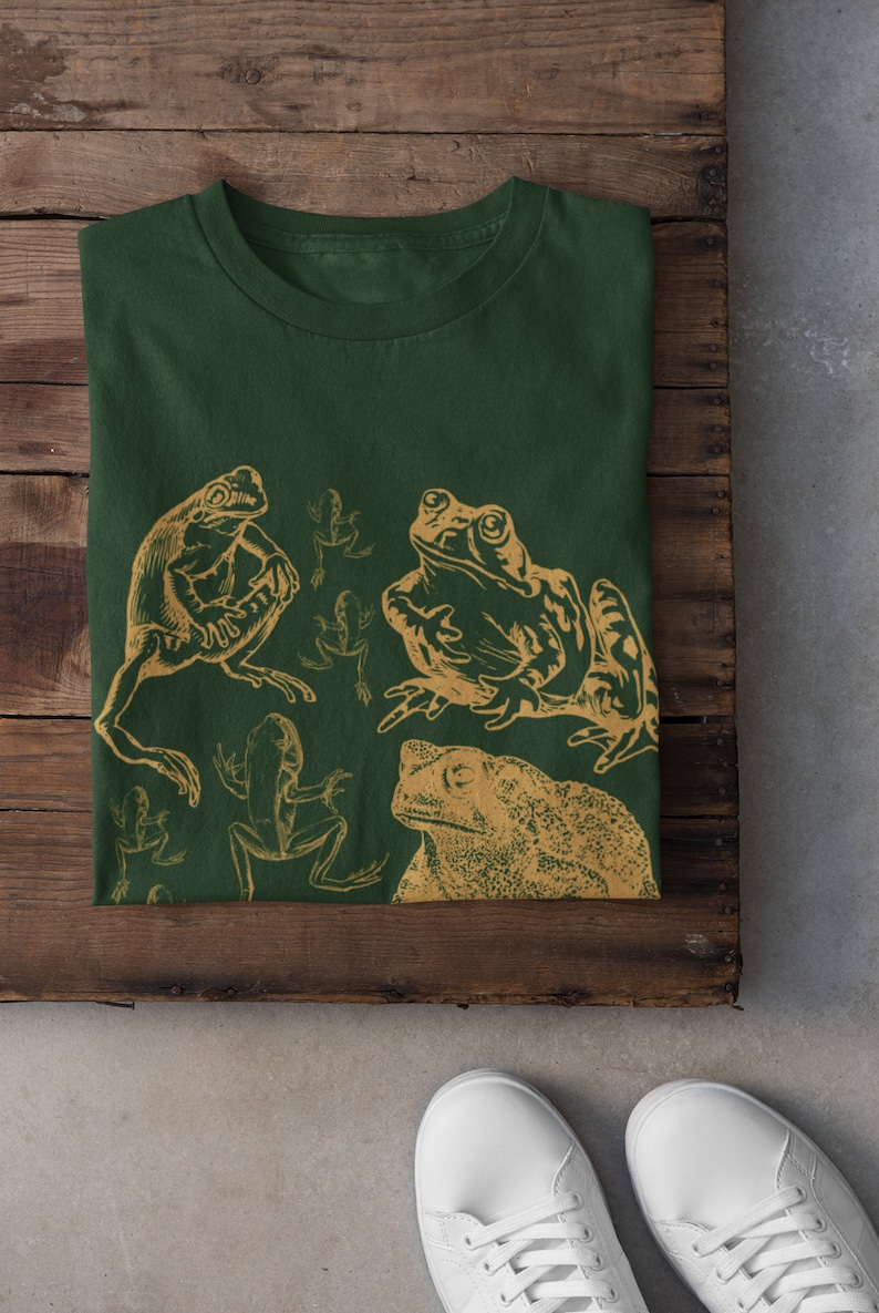 Pode incluir: Uma camiseta verde com uma estampa dourada de sapos e r&atilde;s. A estampa apresenta uma variedade de poses de sapos, incluindo um sapo grande com um olho proeminente.