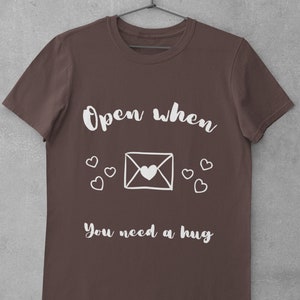 Pode incluir: Camiseta marrom com o texto branco "Open when you need a hug". O design inclui um envelope branco com um coração e vários corações pequenos. A camiseta está pendurada em um cabide de metal.