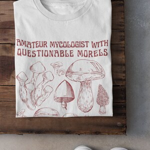 Może przedstawiać: Biała koszulka z czerwonym nadrukiem grzyb&oacute;w i tekstem "Amateur Mycologist with Questionable Morels".