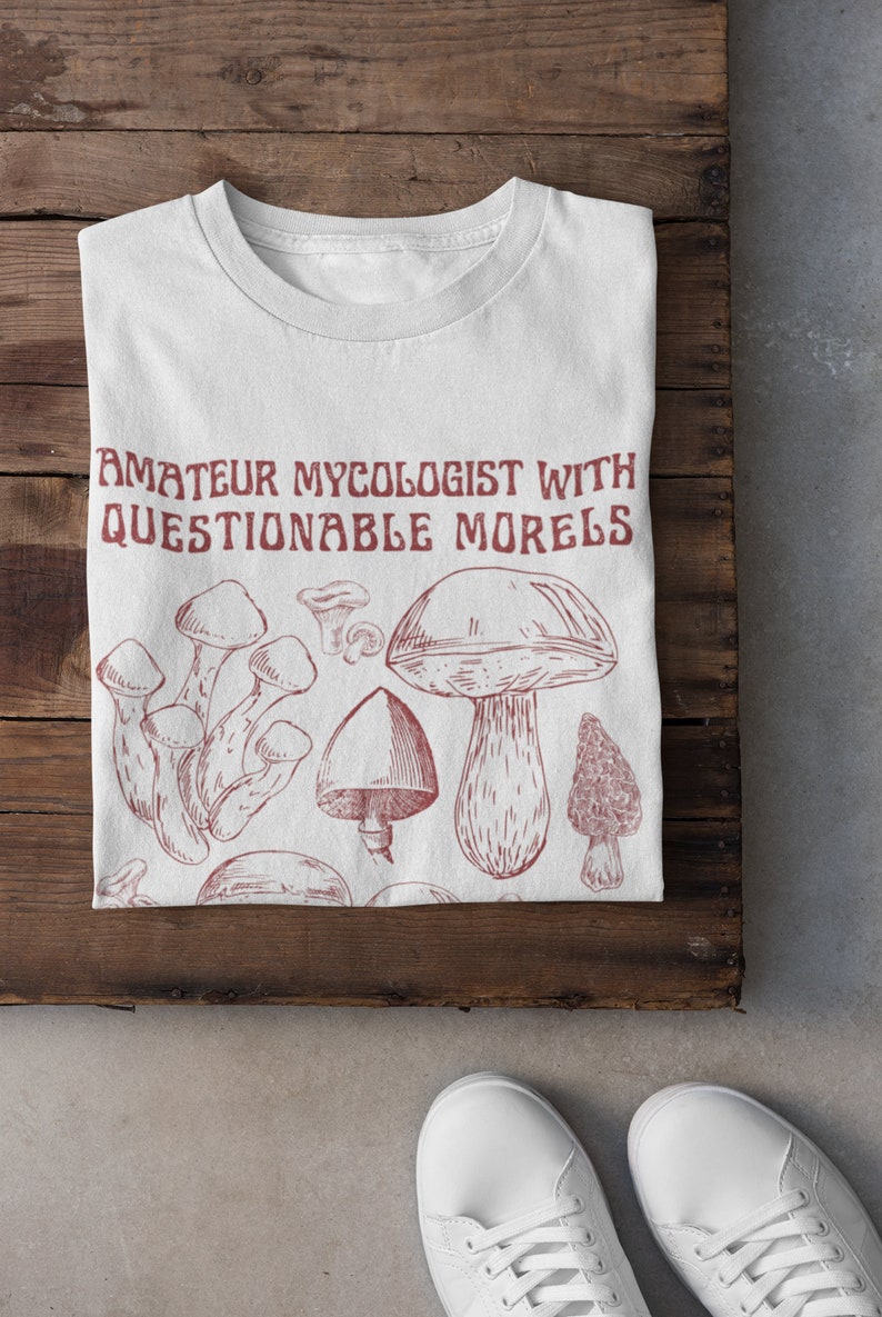 Może przedstawiać: Biała koszulka z czerwonym nadrukiem grzyb&oacute;w i tekstem "Amateur Mycologist with Questionable Morels".