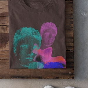 May include: A brown t-shirt with a graphic print of three faces in shades of pink, blue, and green. The faces are stylized and appear to be from classical sculptures.
