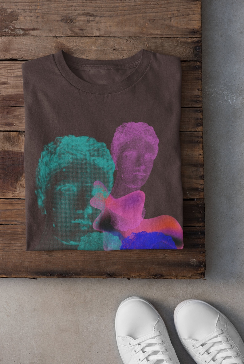 May include: A brown t-shirt with a graphic print of three faces in shades of pink, blue, and green. The faces are stylized and appear to be from classical sculptures.