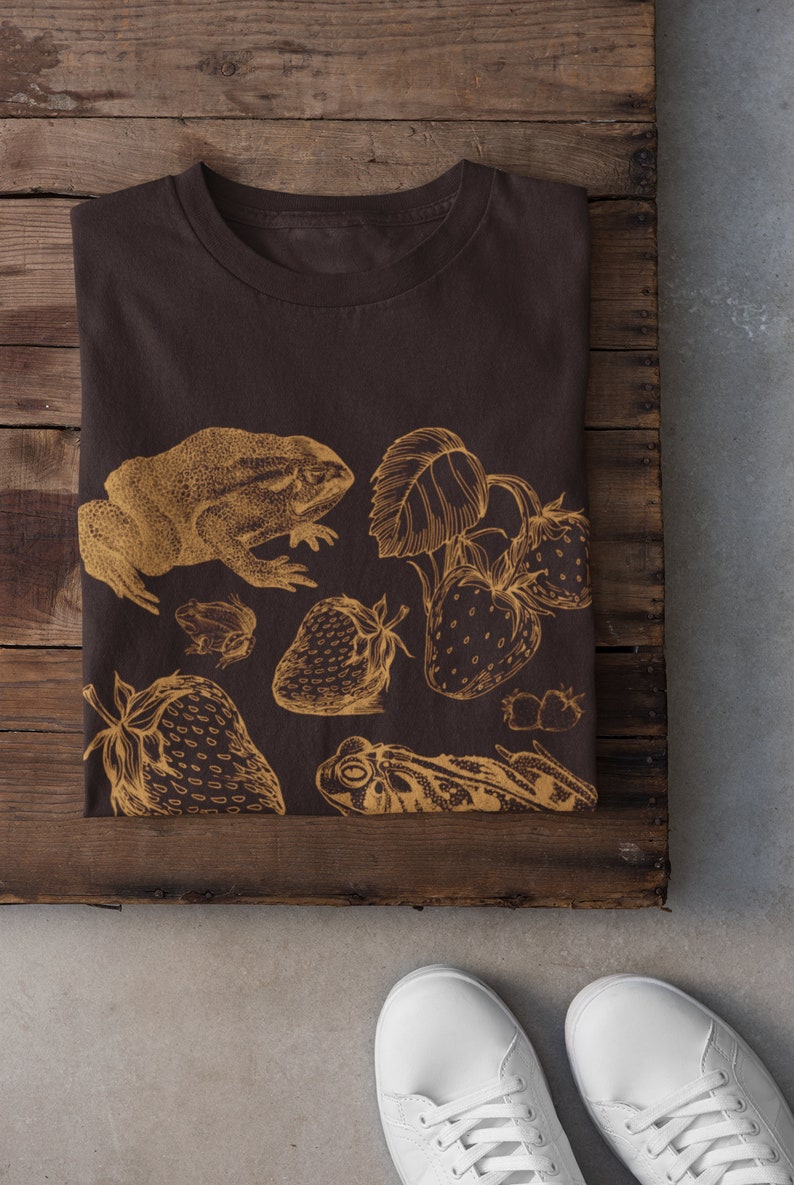 Pu&ograve; includere: Una maglietta marrone con una stampa dorata di rane e fragole. La stampa &egrave; un'illustrazione in stile vintage.