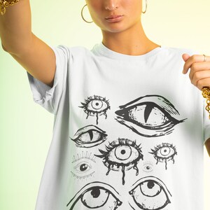 May include: White t-shirt with a graphic print of multiple black and white eyes. The eyes are drawn in a stylized, abstract style.