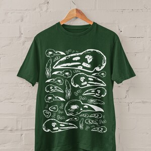 Op de afbeelding: Donkergroen t-shirt met een witte afbeelding van vogel schedel illustraties. Het ontwerp toont schedels in verschillende maten en vormen, die een herhalend patroon vormen. Het t-shirt hangt aan een houten hanger tegen een witte bakstenen muur.
