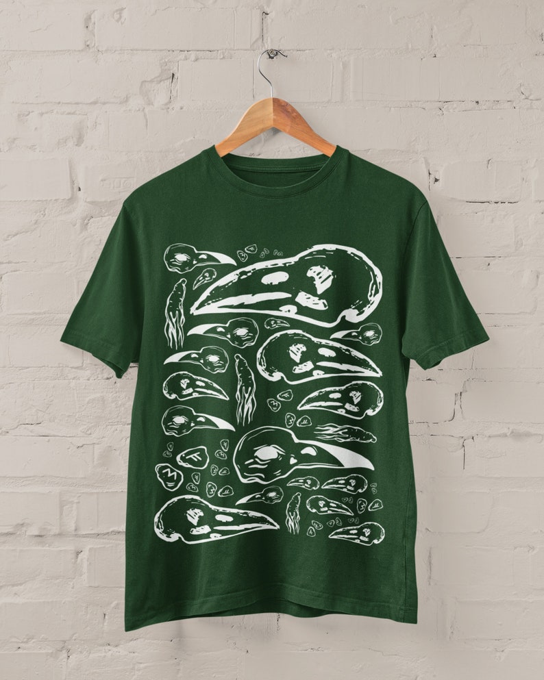 Op de afbeelding: Donkergroen t-shirt met een witte afbeelding van vogel schedel illustraties. Het ontwerp toont schedels in verschillende maten en vormen, die een herhalend patroon vormen. Het t-shirt hangt aan een houten hanger tegen een witte bakstenen muur.