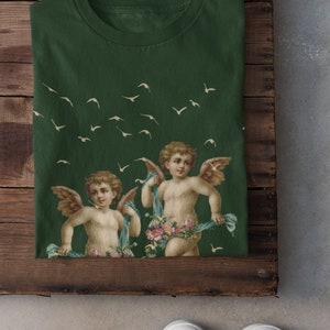 May include: A green t-shirt with a print of two cherubs with wings and flowers. The cherubs are holding blue ribbons. There are white birds flying in the background.