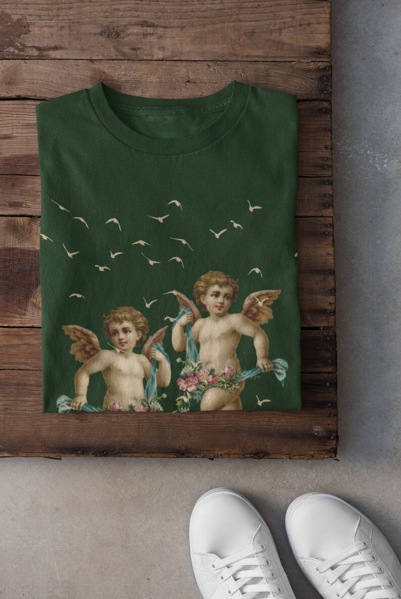 May include: A green t-shirt with a print of two cherubs with wings and flowers. The cherubs are holding blue ribbons. There are white birds flying in the background.